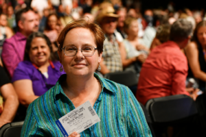 Woman with short hair and glasses, wearing a teal striped shirt, sitting in a crowded audience and holding a ticket or pass at chest level.