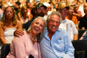 Joyful elderly couple posing for the camera with arms around each other at a crowded event auditorium.