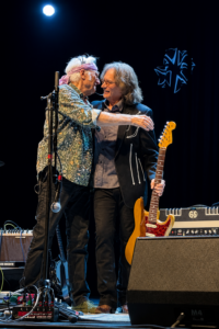 Two male musicians share a hug on stage, with a yellow electric guitar and gear nearby.