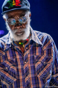 Close-up portrait of an elderly man with a white beard, wearing a cap with a red dragon embroidery, round glasses, and a multicolored bead necklace over a plaid shirt.