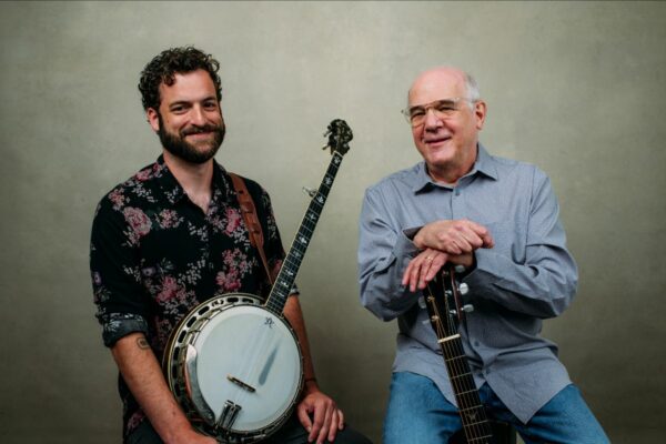 Two men pose with musical instruments: a banjo on the left and a guitar on the right, both smiling at the camera.