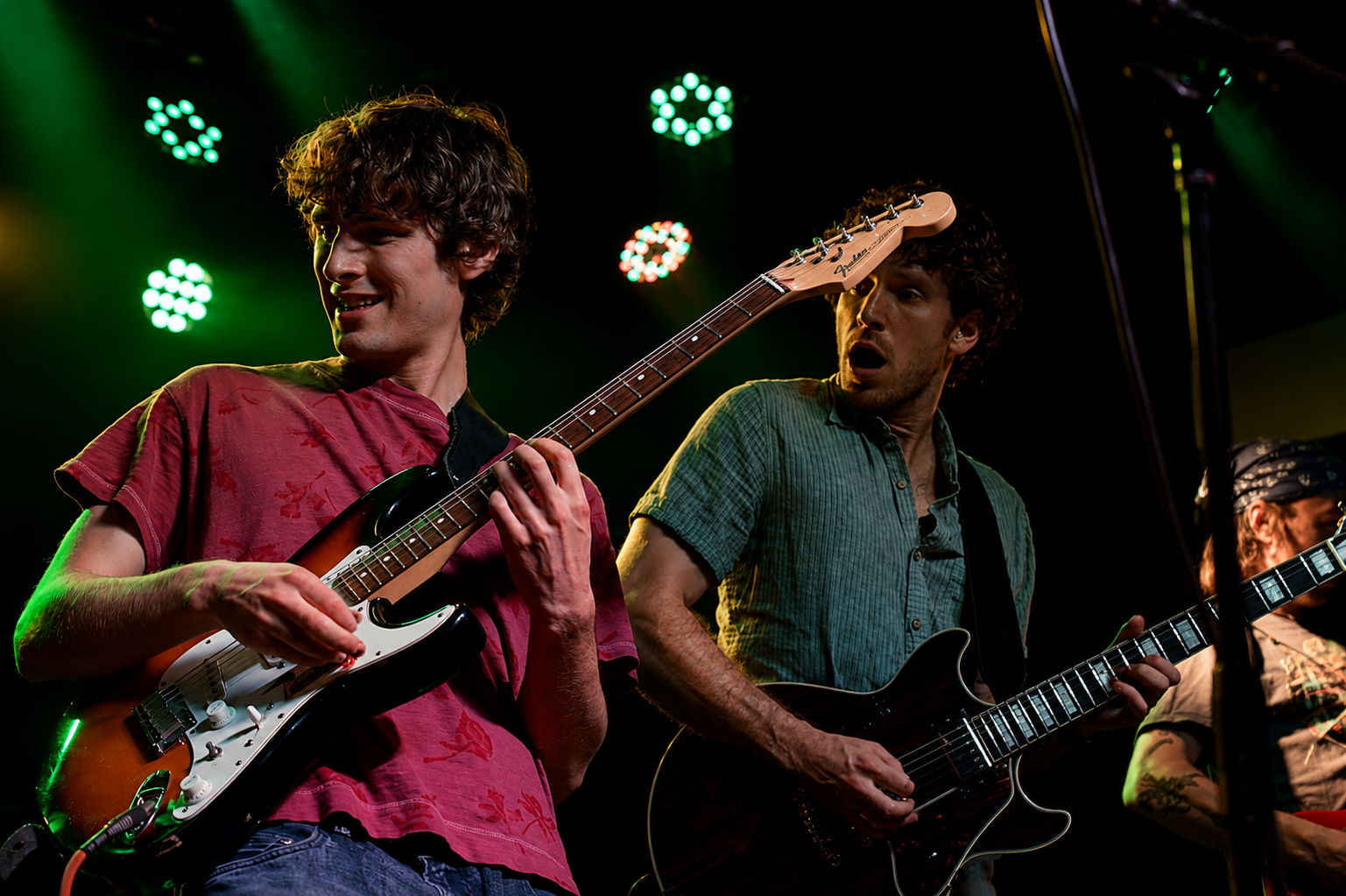 Two male guitarists perform on stage under green lights, playing electric guitars.
