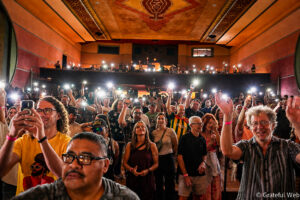 Crowd at a concert in a theater, many holding up phone flashlights, with a balcony and ornate ceiling above.