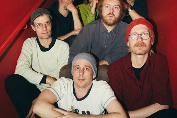 Six friends sit on a red staircase, posing for the camera with relaxed, neutral expressions in casual clothing.