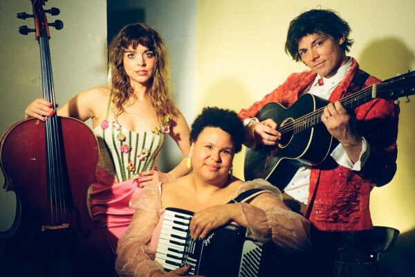 Three musicians posing with instruments: cello, accordion, and guitar in a studio setting.