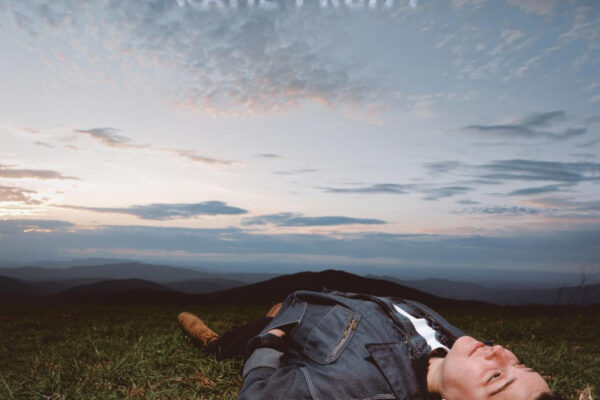Person lying on grass on a hillside at sunset, wearing a denim jacket, with distant mountains and a blue sky above.