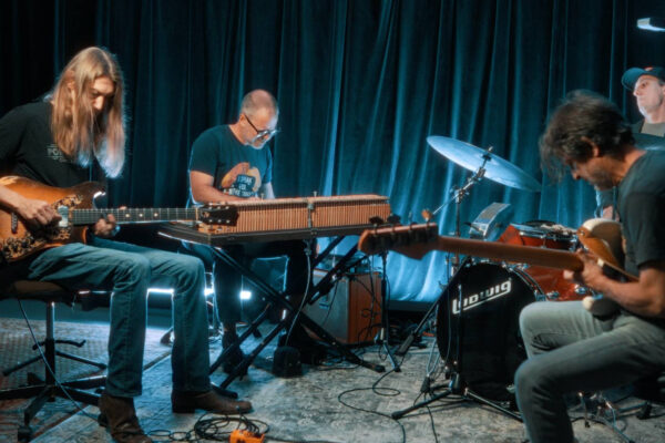 Band rehearsal with four musicians: guitarist left, keyboard in center, drummer on right, and another guitarist in foreground on stage