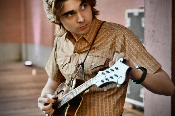 Young man playing an acoustic guitar outdoors, wearing a brown patterned shirt and leather belt.