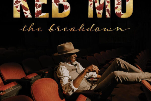 Man in a fedora lounging in red theater seats under a large gold-text sign that reads 'Keb' Mo' the breakdown'.