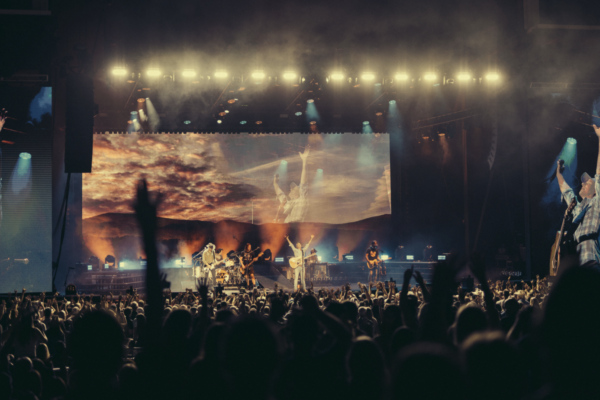 Wide view of a live concert with a rock band performing on stage, screens on left and right showing performers, and a crowd raising hands in front of the stage, dark venue with bright lights above.