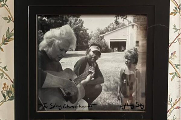 Black-and-white framed photo of three people outdoors; an older person strums a guitar while a man and a young girl stand nearby, with a barn in the background. The image includes handwritten captions: 'The String Cheese' and 'Lightin(g) Sky'.