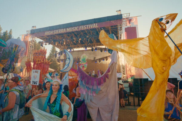Colorful festival parade in front of a stage labeled High Sierra Music Festival, performers in flowing winged costumes.