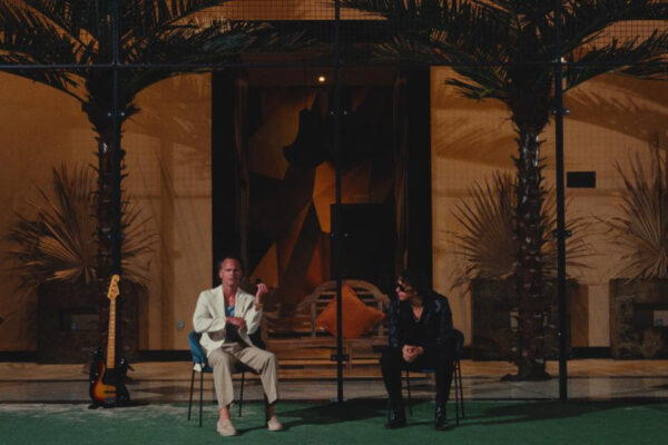 Two men sit on chairs outdoors at night, chatting with a bass guitar and a conga drum nearby, in a tropical-themed venue.
