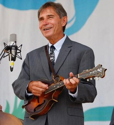 Smiling man in a gray suit playing a mandolin on stage beside a microphone.