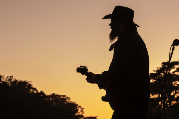 Silhouette of a guitarist wearing a cowboy hat performing at sunset with a microphone nearby.