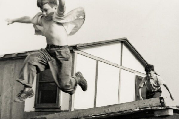 Two boys play on a rooftop: one shirtless in mid‑jump with a cape-like shirt fluttering behind him; the other boy stands nearby on the roof edge.