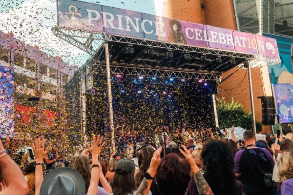 Crowd cheers at an outdoor stage for a 'Prince Celebration' with confetti raining down from above.