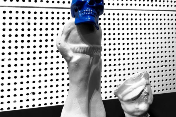 Tall white sculpture with a bright blue skull for a head, placed on a black surface against a dotted wall.
