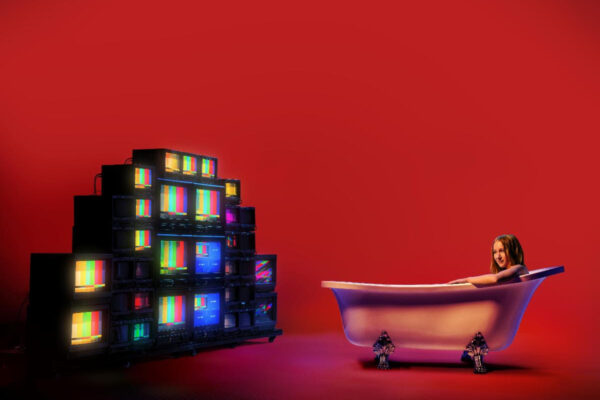 Girl sits in a white clawfoot bathtub while a stack of colorful CRT televisions glows beside her on a red backdrop.