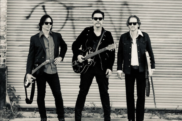 Three male rock musicians with guitars pose in front of a graffiti-marked corrugated metal wall, wearing sunglasses.