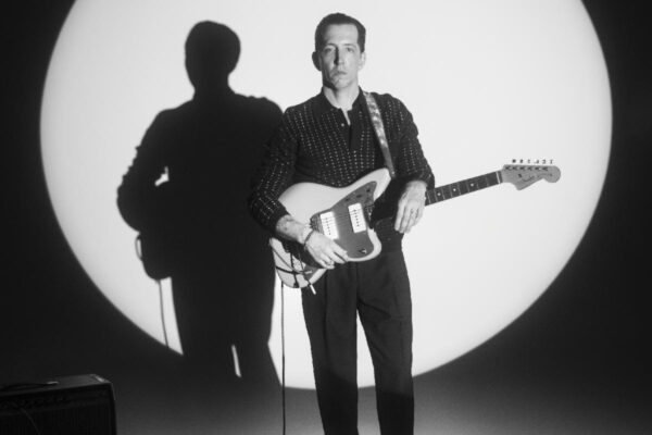 Guitarist standing on stage in front of a large circular spotlight, holding an electric guitar.
